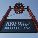 Signage of the Science and Industry Museum in Manchester, featuring a red gear design against a clear blue sky